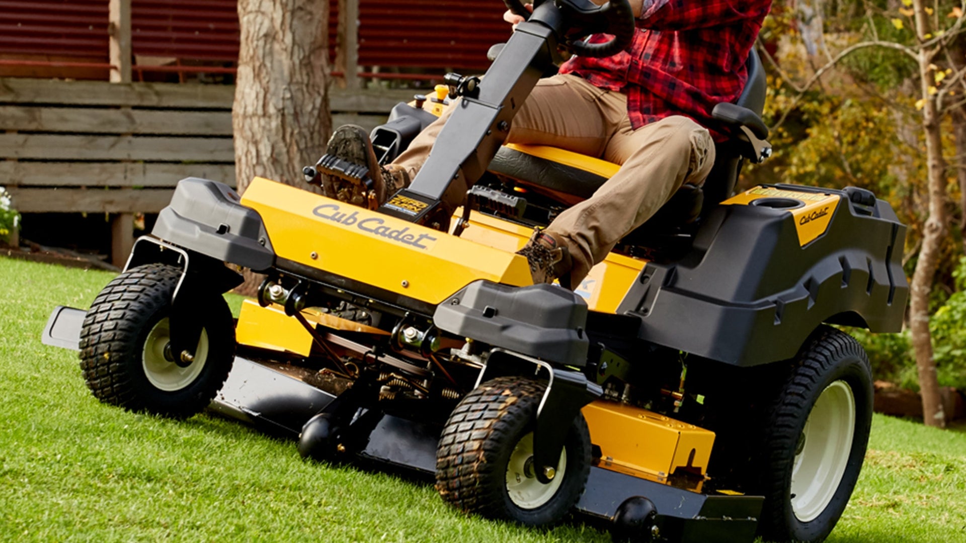 Main in checkered shirt steering his yellow Cub Cadet ride on mower across lawn with trees in background.