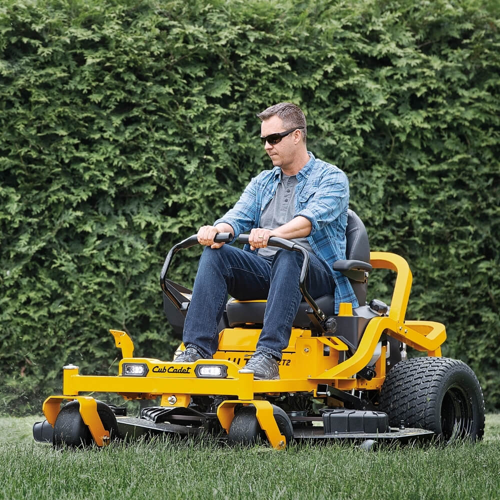 Man operating a yellow Cub Cadet lawn mower in a grassy area with green bushes in the background