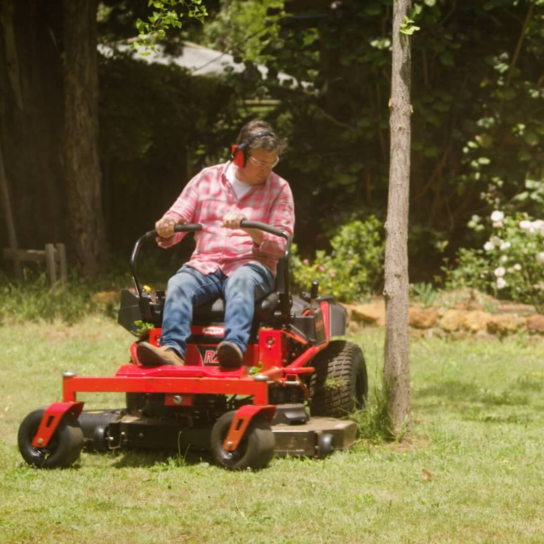 Man wearing ear protection navigating his red ride on mower around a tree.