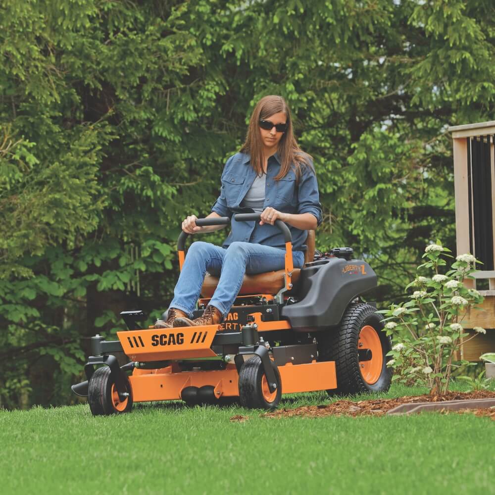 Woman wearing glasses operating a SCAG Liberty riding lawn mower in a garden setting
