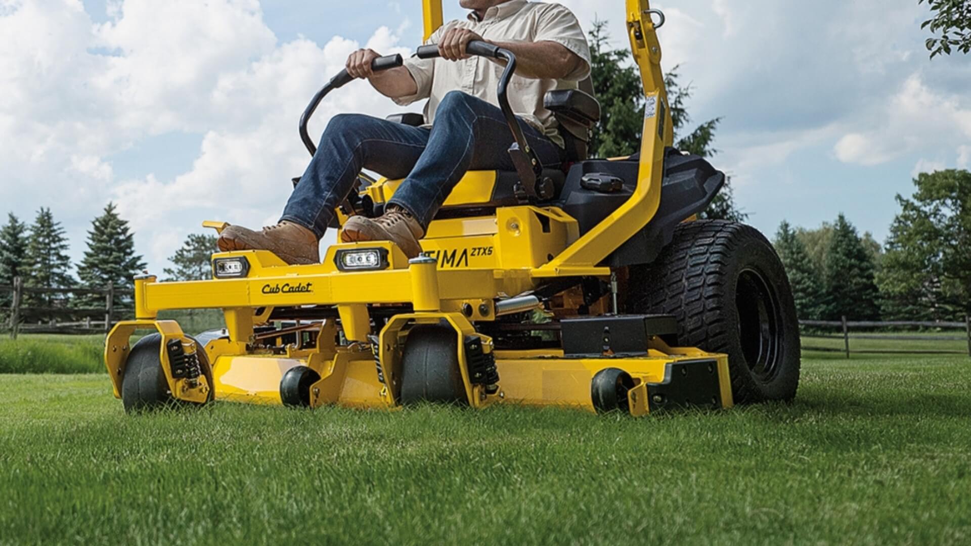 Person operating a yellow stand-on lawn mower in a grassy area with trees in the background.