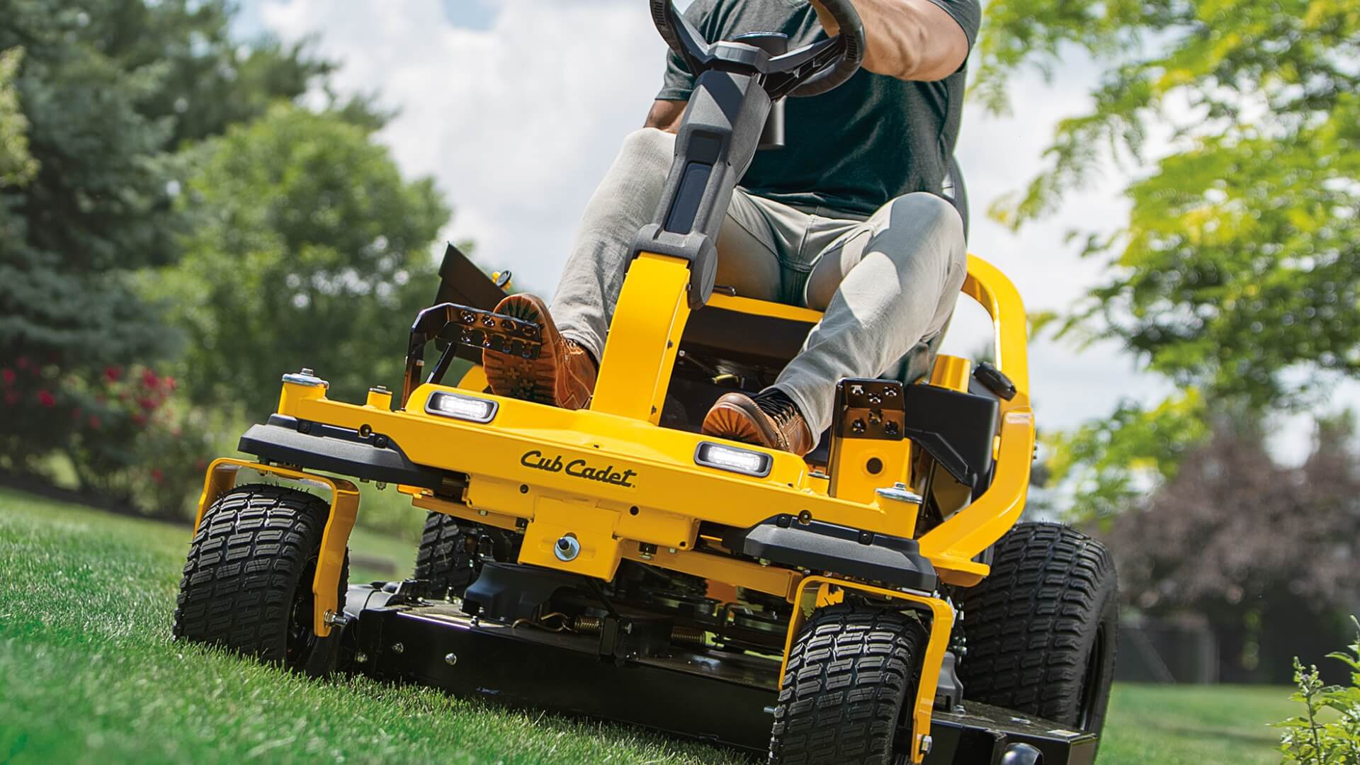 Person operating a yellow Cub Cadet lawn mower on a grassy area with trees in the background.