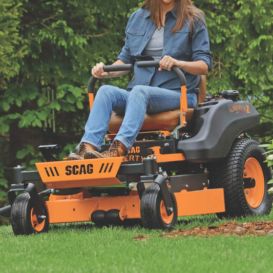 Person operating an orange SCAG lawn mower in a grassy area with trees in the background