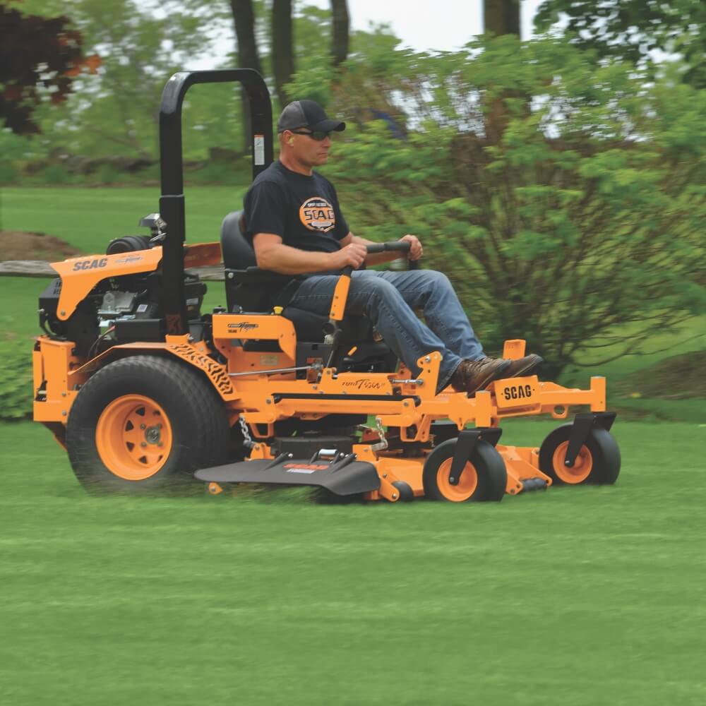 Person operating a SCAG riding lawn mower on a grassy area with trees in the background