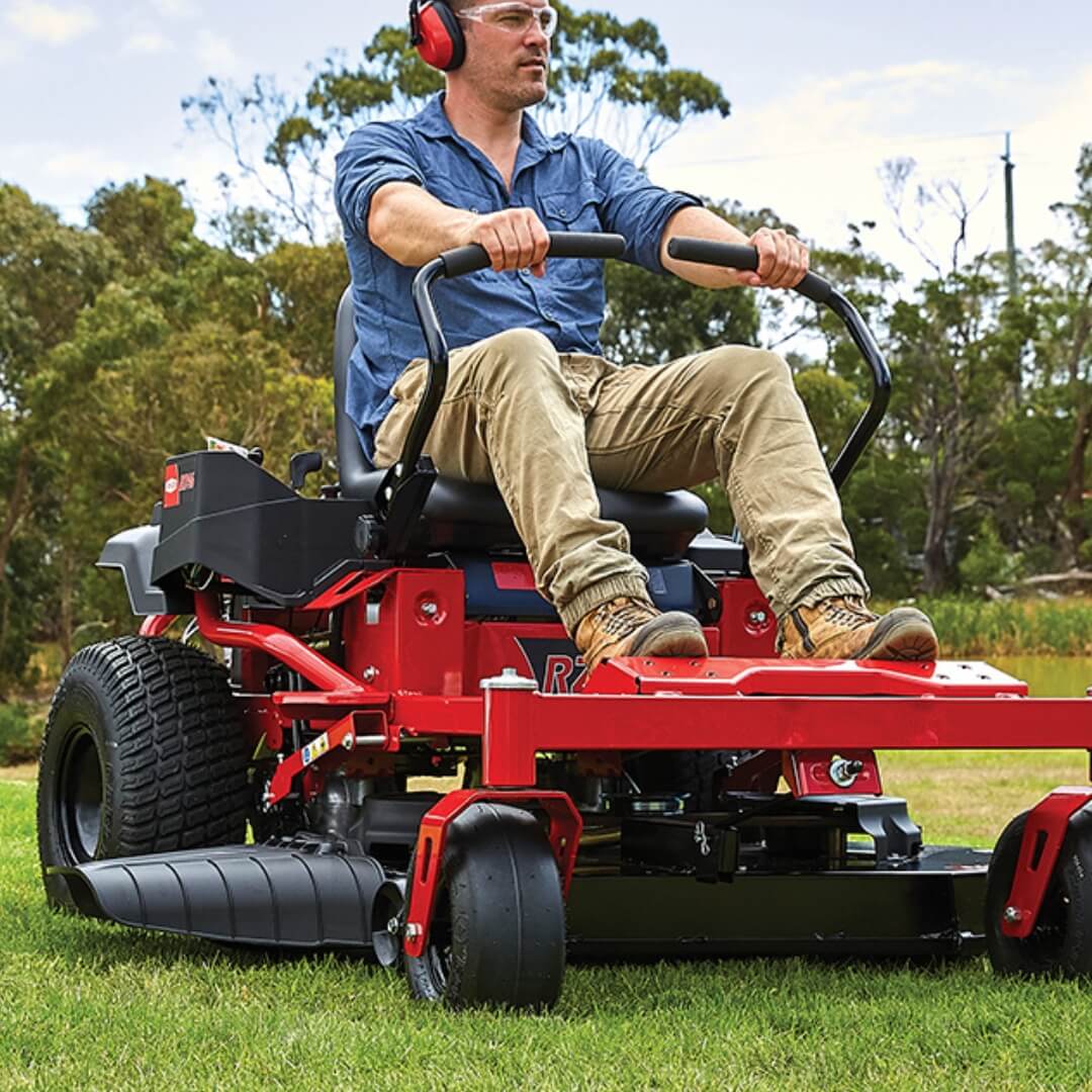 Man operating a red riding lawn mower in a grassy area with trees in the background