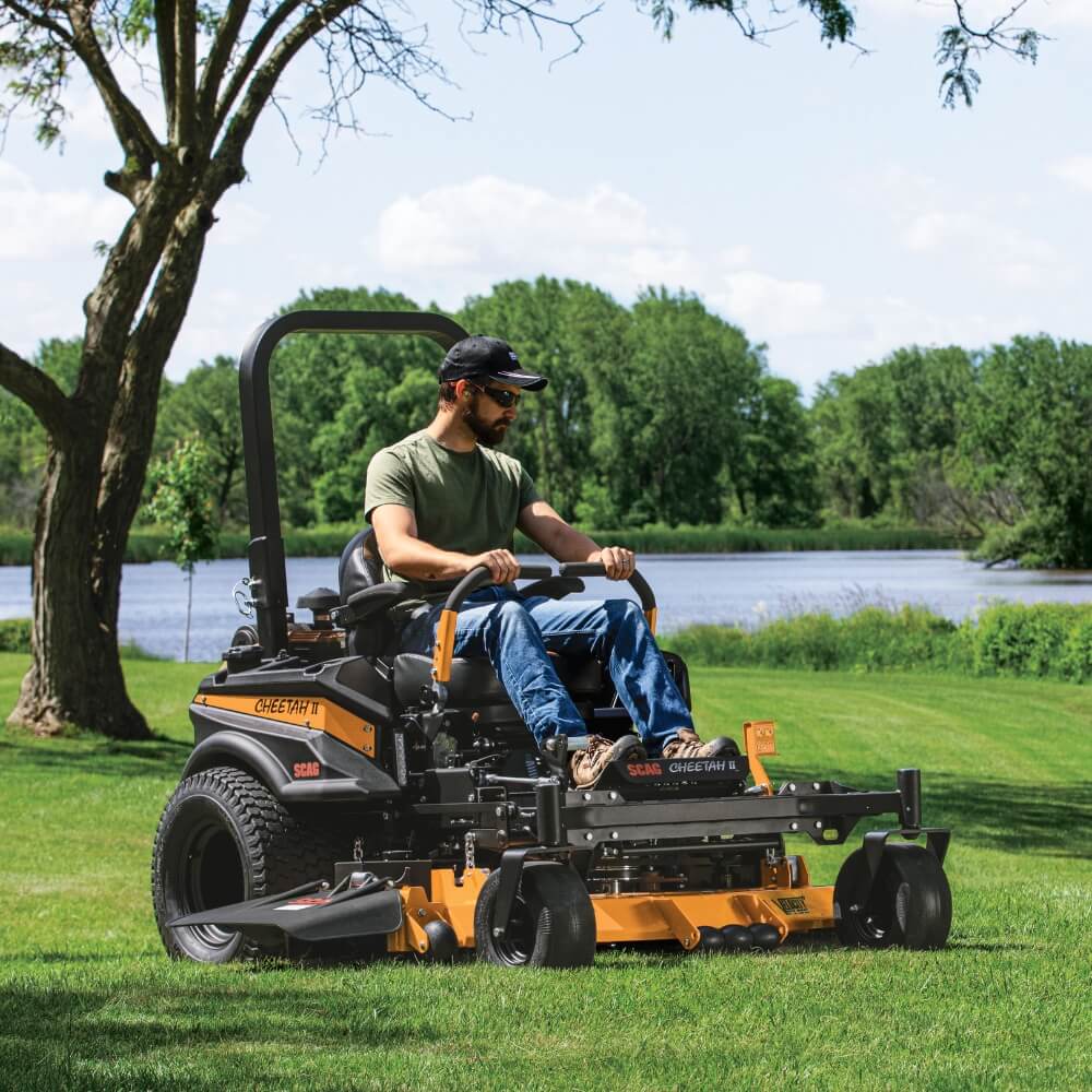 Man operating a SCAG Cheetah riding lawn mower in a park with trees and water in the background